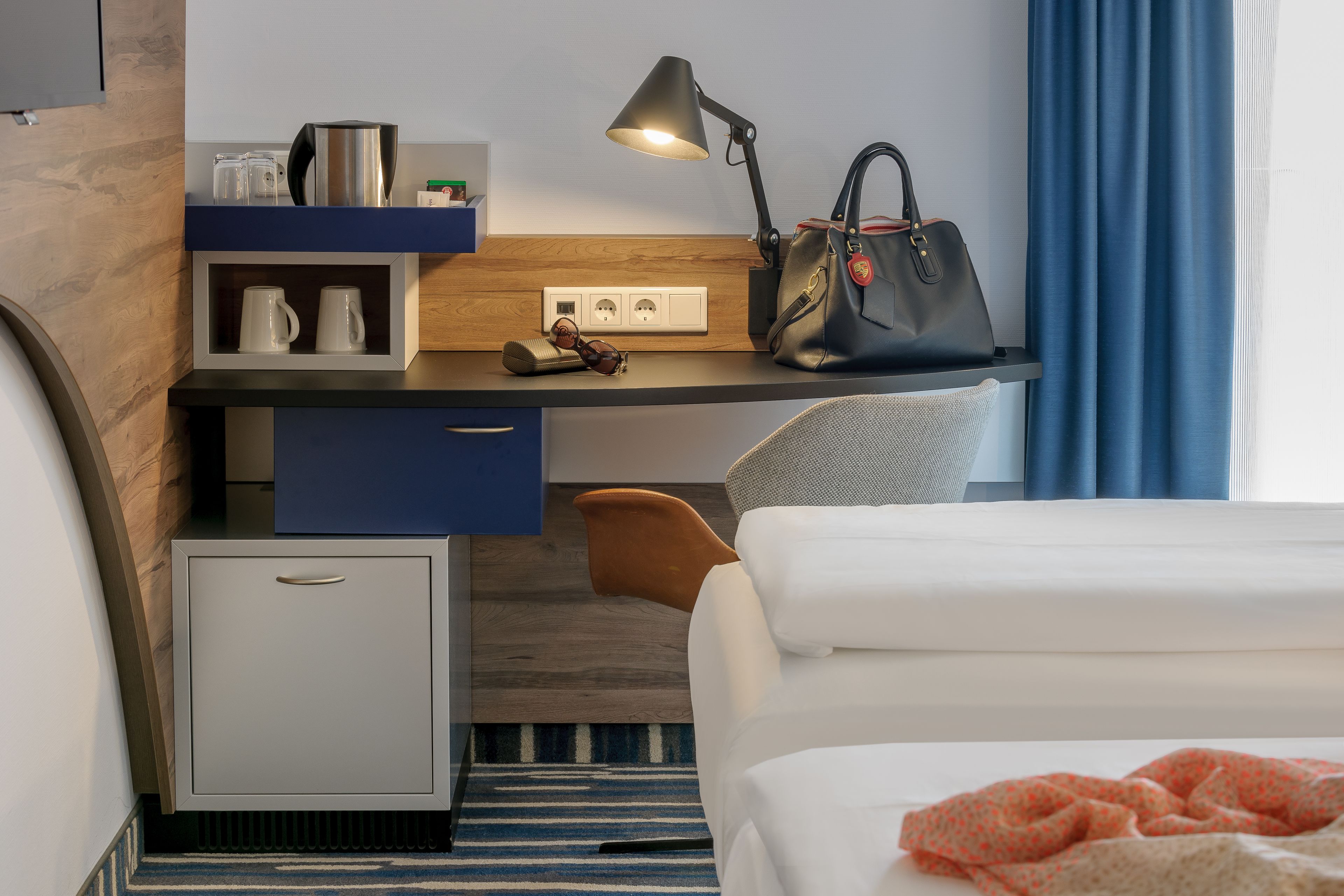 A hotel room with a bed, a desk with a lamp, a handbag, and a small fridge.