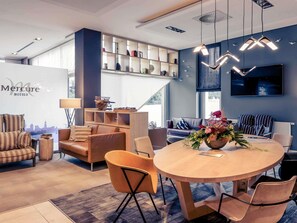 Moderni hotellihuone, jossa on ruokailutila, sohvaryhmä, seinälle asennettu televisio ja kirjahylly, jossa on erilaisia esineitä.