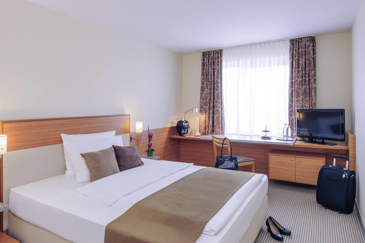 Una habitación de hotel con cama, un escritorio con televisor, una silla, una maleta y una ventana con cortinas.