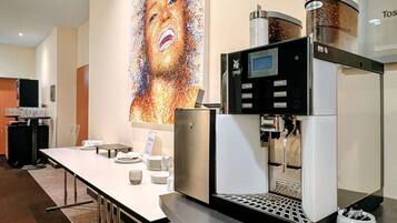 Una máquina de café moderna, una mesa larga con platos y vasos, y un cuadro abstracto grande en la pared.
