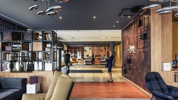 Un hall d’hôtel moderne comprenant un comptoir d’accueil, un espace salon avec des canapés, une bibliothèque et un mur décoratif en bois.