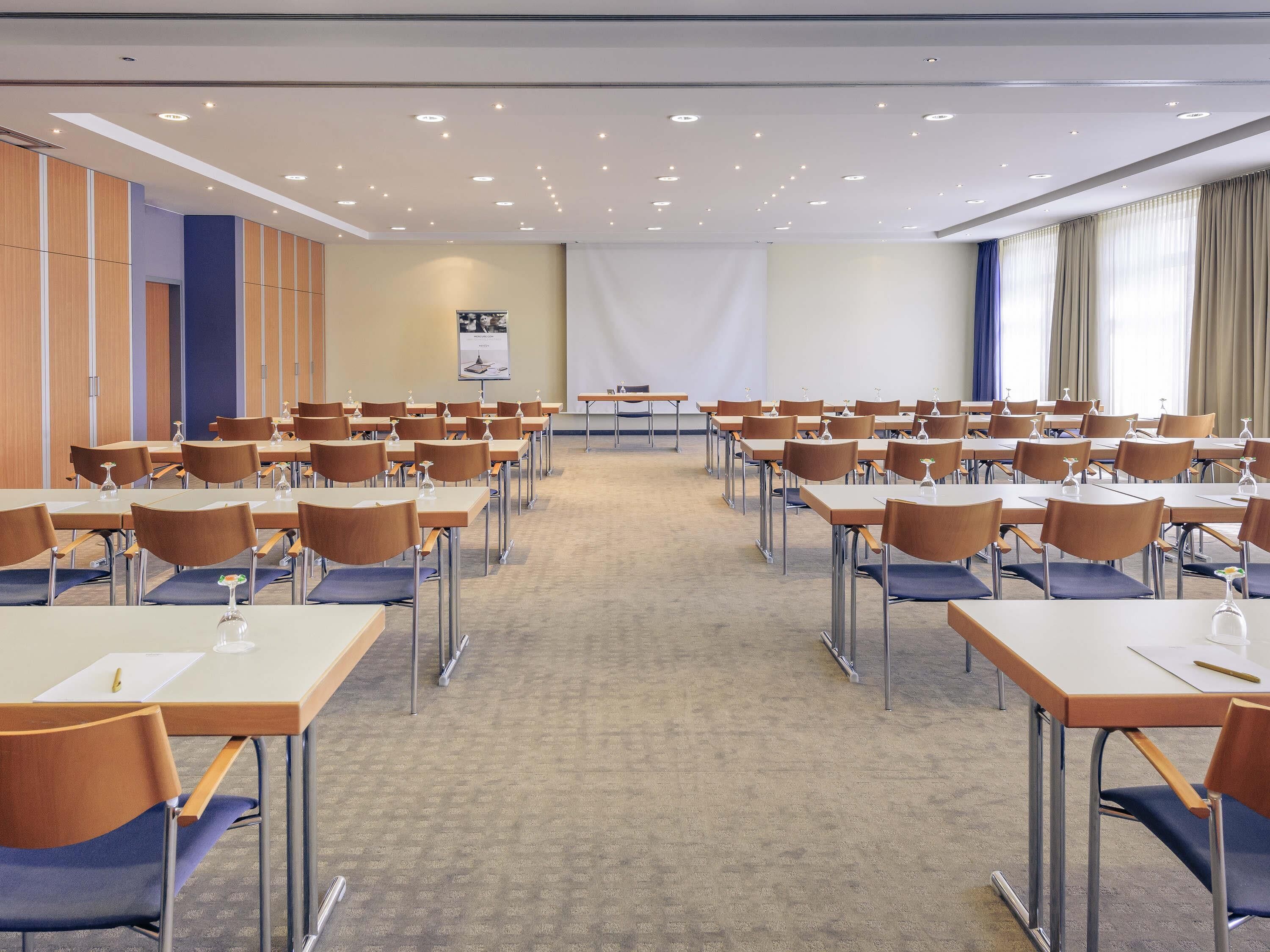A conference room with rows of tables and chairs, a projector screen, and a large window with curtains.