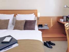 Quarto de hotel com uma cama bem arrumada, uma mesa de cabeceira com abajur, uma pequena estante com cafeteira e um par de sapatos no chão.