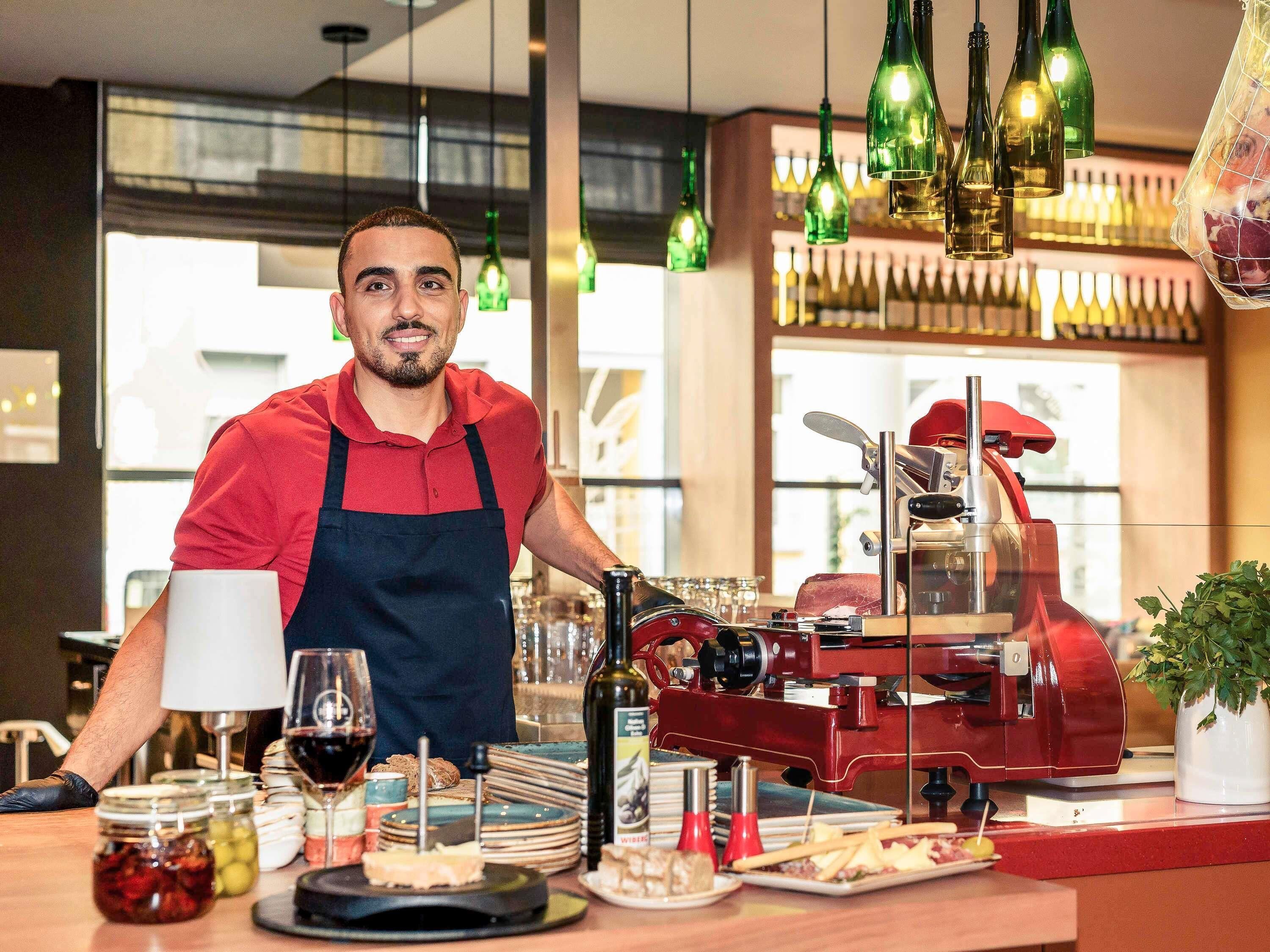 A restaurant interior with a chef preparing food, a meat slicer, and various food items on the counter.