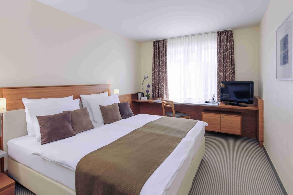 A hotel room with a bed, a desk with a TV, a chair, and a lamp.