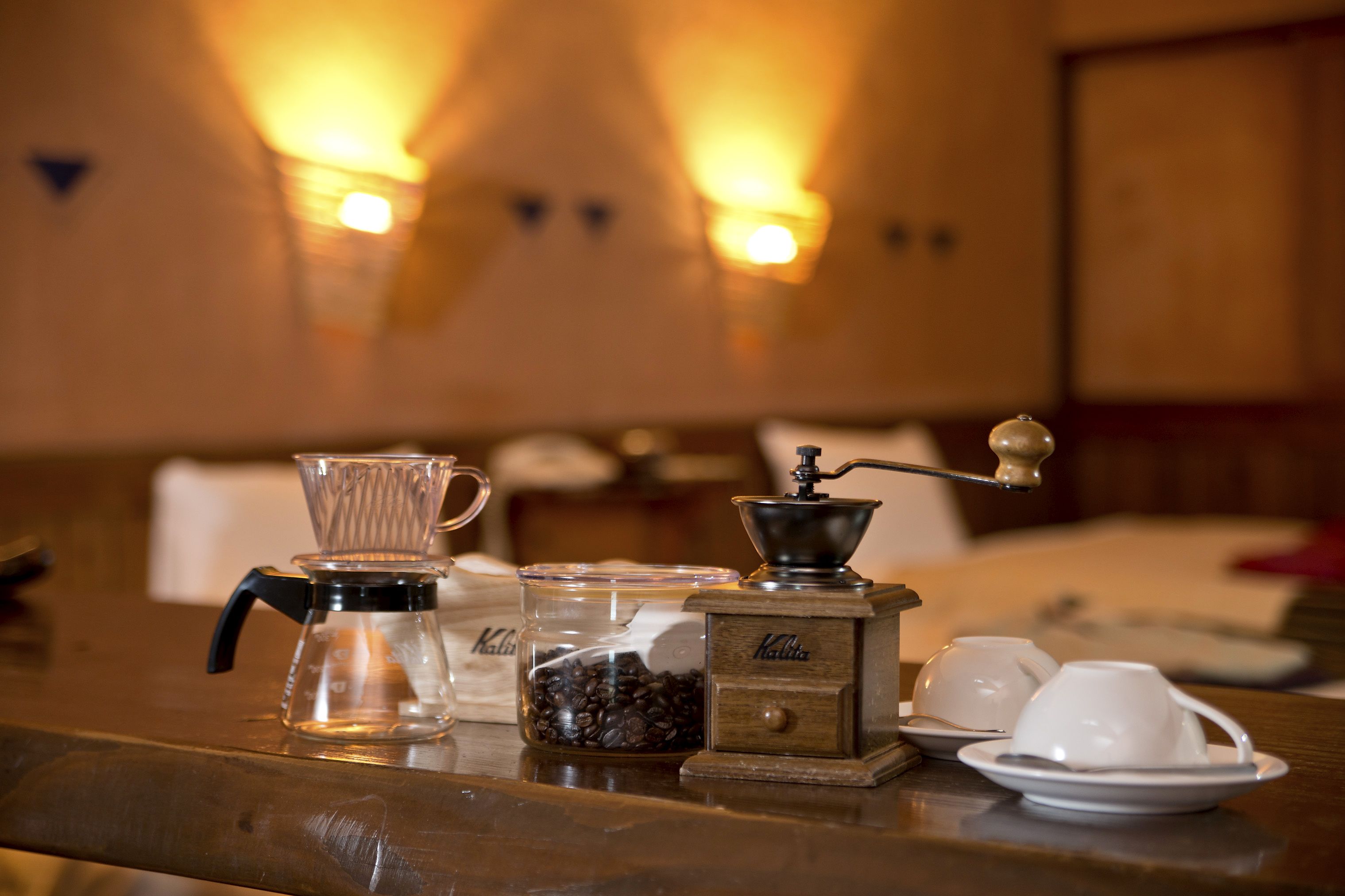 A coffee station with a coffee grinder, a coffee pot, a glass container with coffee beans, and two white cups on a wooden table.