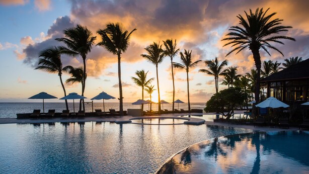 Zona de piscina con sillones de descanso y sombrillas, palmeras y vistas al atardecer.