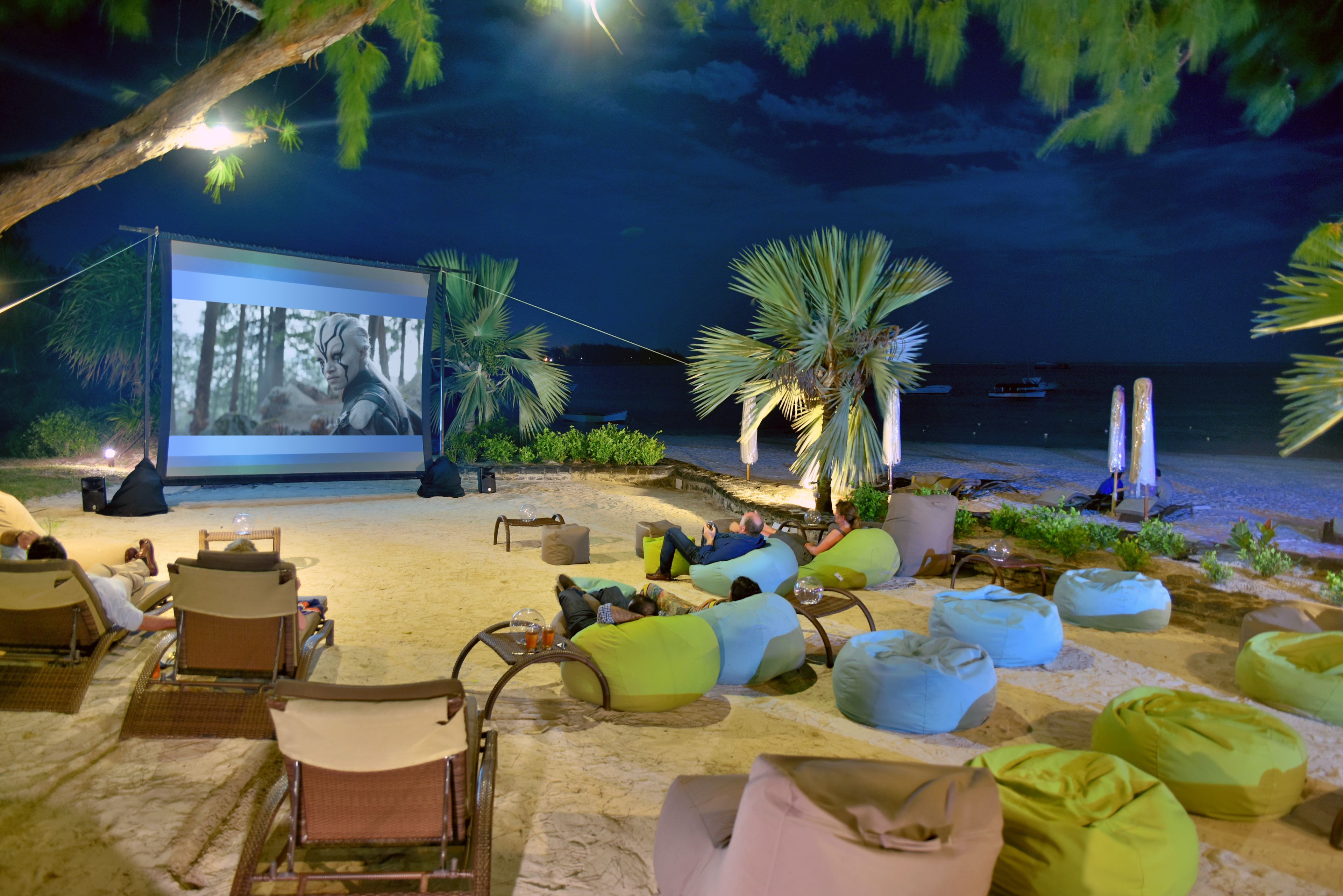 An outdoor cinema with bean bag seating and a large screen, set on a sandy beach with palm trees and a view of the sea.