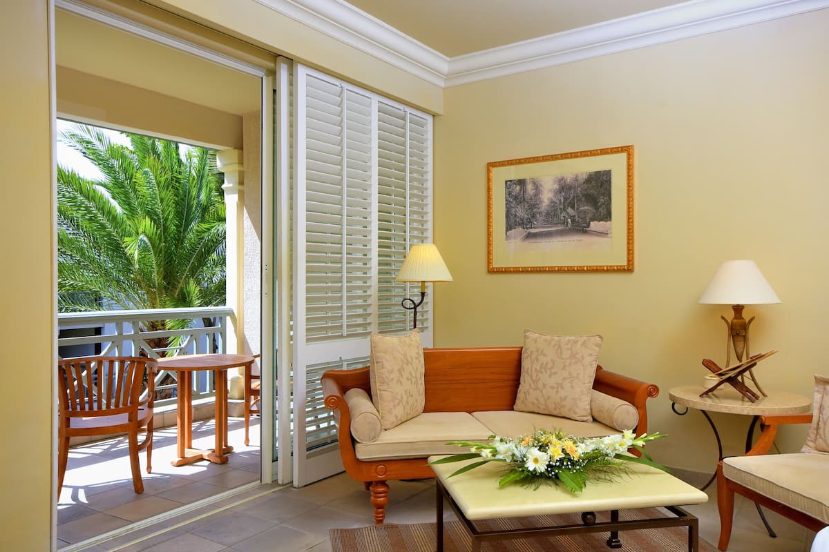 A room with a wooden sofa, a small table with a flower arrangement, a chair, and a lamp. There is a window with blinds and a picture on the wall.