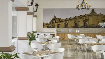 Un comedor moderno con sillas y mesas blancas, una araña y un mural de un edificio en la pared.