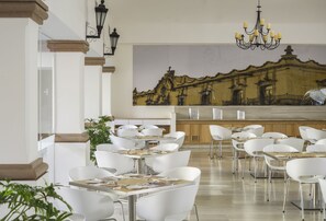Un espace de restauration moderne avec des chaises et des tables blanches, un lustre et une fresque représentant un bâtiment sur le mur.