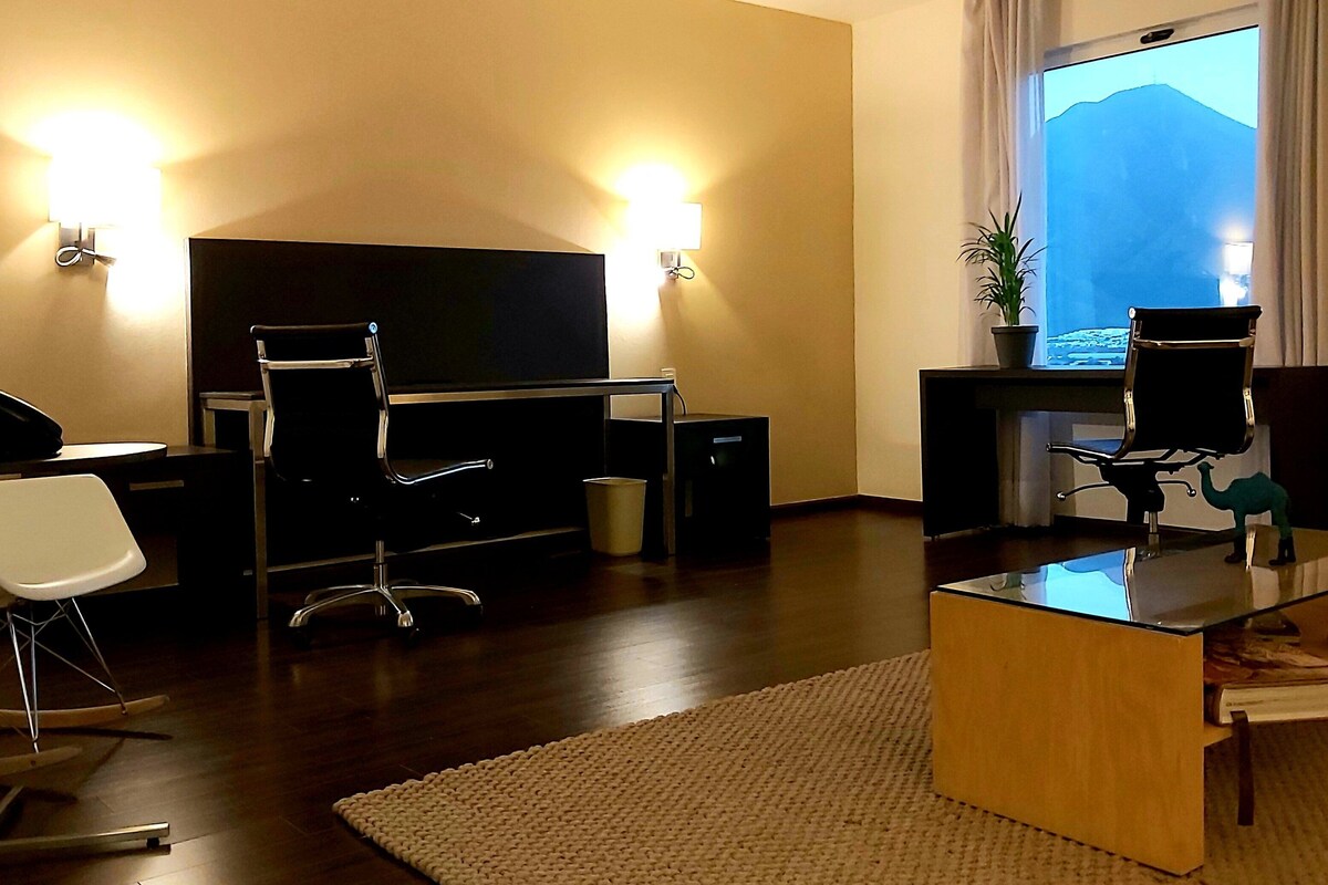 Habitación de hotel moderna con televisión de pantalla plana, un escritorio con computadora, una silla blanca, una mesita con planta y vistas a las montañas desde la ventana.