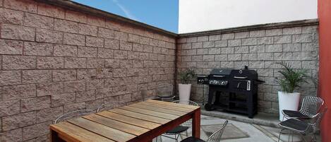 A patio area with a wooden table and metal chairs, a barbecue grill, and potted plants.
