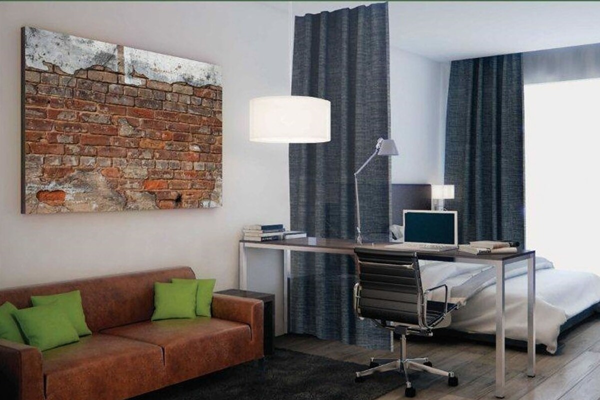 Una habitación de hotel con un mural de ladrillos, un escritorio con una computadora, una cama, un sofá y una ventana con cortinas.