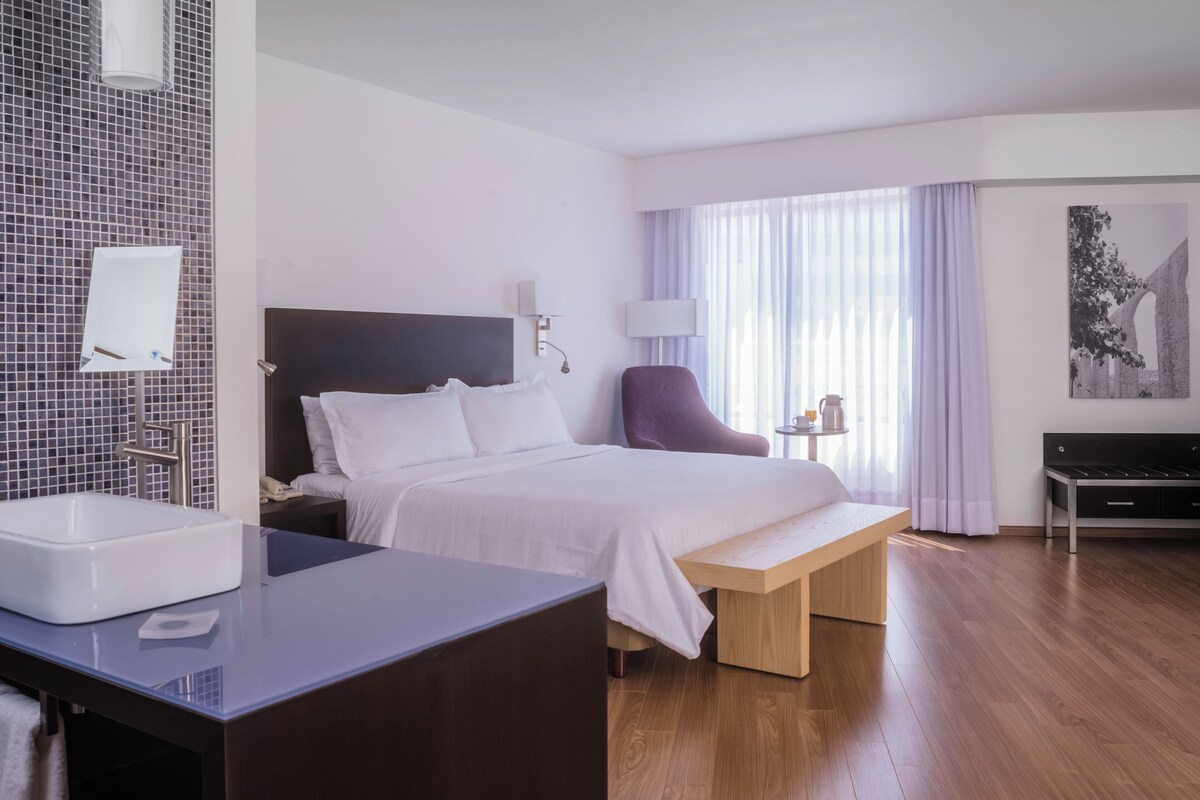 Habitación de hotel con cama, mesita de noche, lámpara, silla y vistas al exterior a través de amplias ventanas.