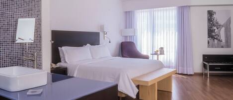 A hotel room with a bed, bedside table, lamp, chair, and a view of the outside through large windows.