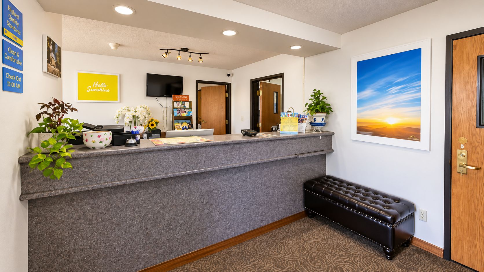 A reception area with a grey reception desk, a bench, a wall-mounted TV, and a framed painting of a sunset.