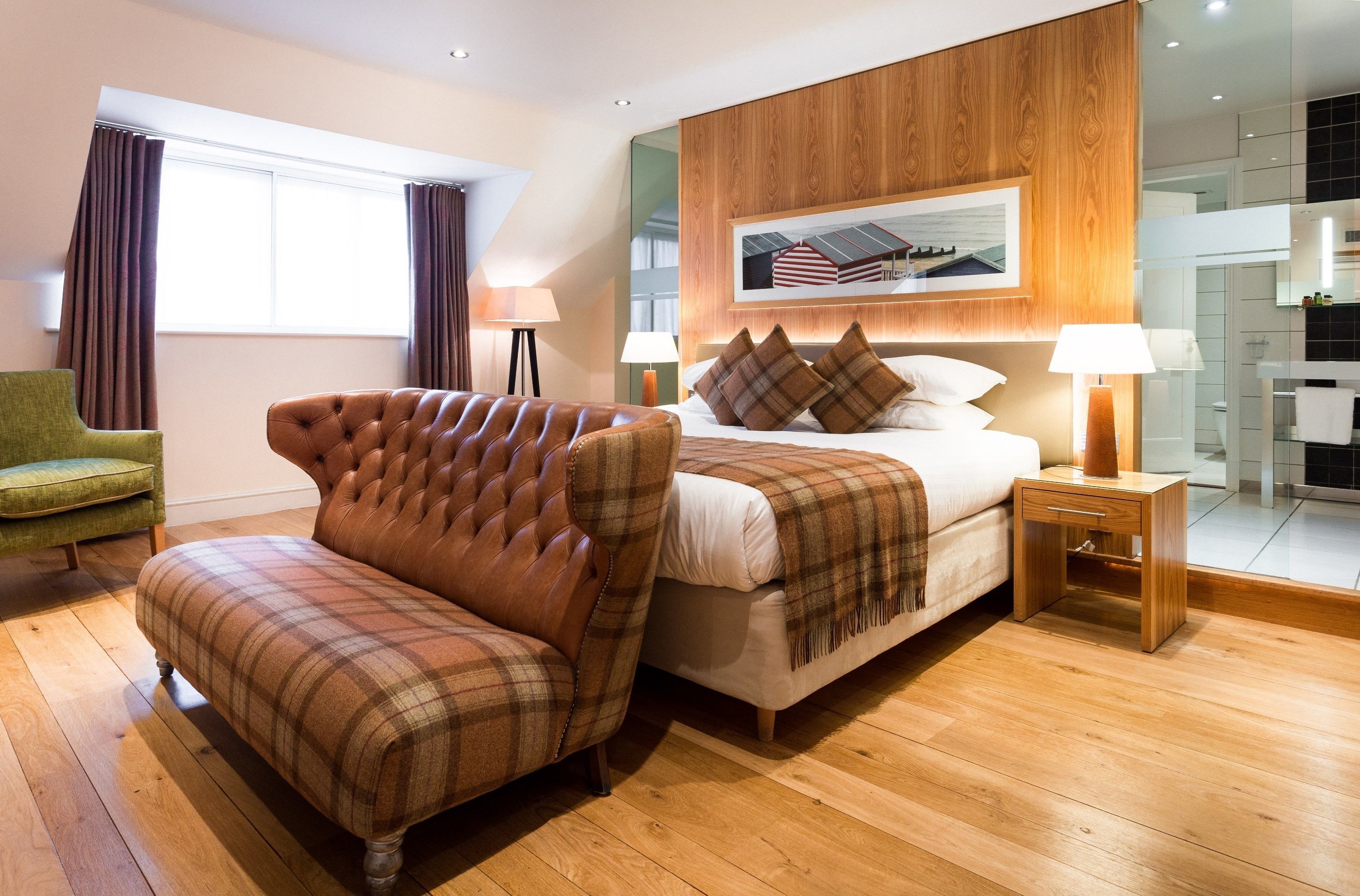 A modern hotel room with a wooden floor, a large bed with a headboard, a green armchair, and a view of the bathroom through a glass door.