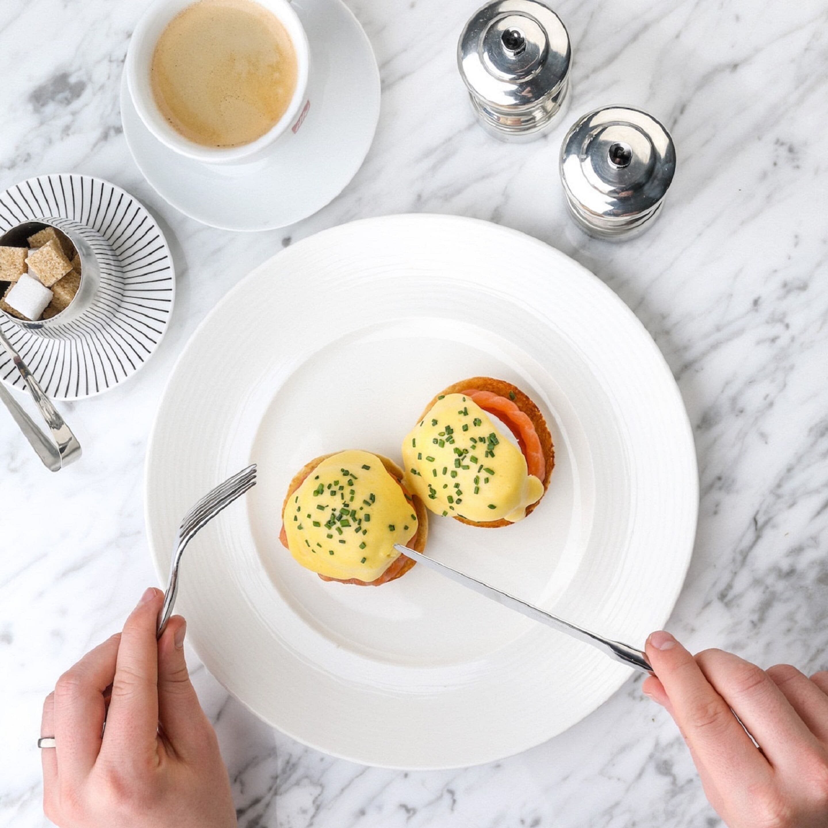 A breakfast setting with two eggs benedict on a white plate, a cup of coffee, and a salt and pepper shaker on a marble surface.