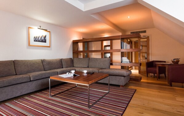 A modern living room with a sectional sofa, a wooden coffee table, and a striped rug. There is a bookshelf with books and a framed picture on the wall.
