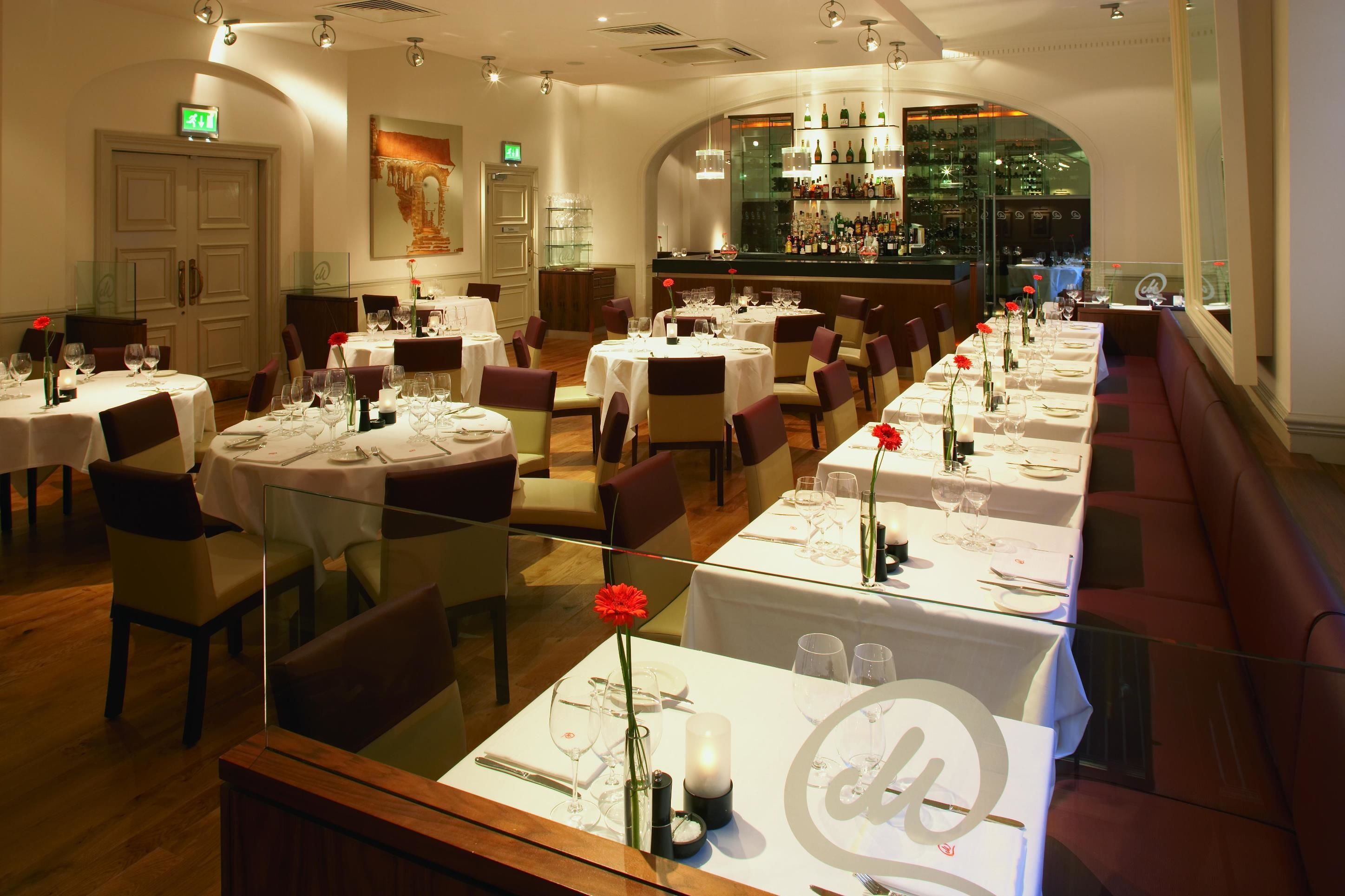 A restaurant with round tables, white tablecloths, and red flowers. There is a bar area with shelves of bottles.