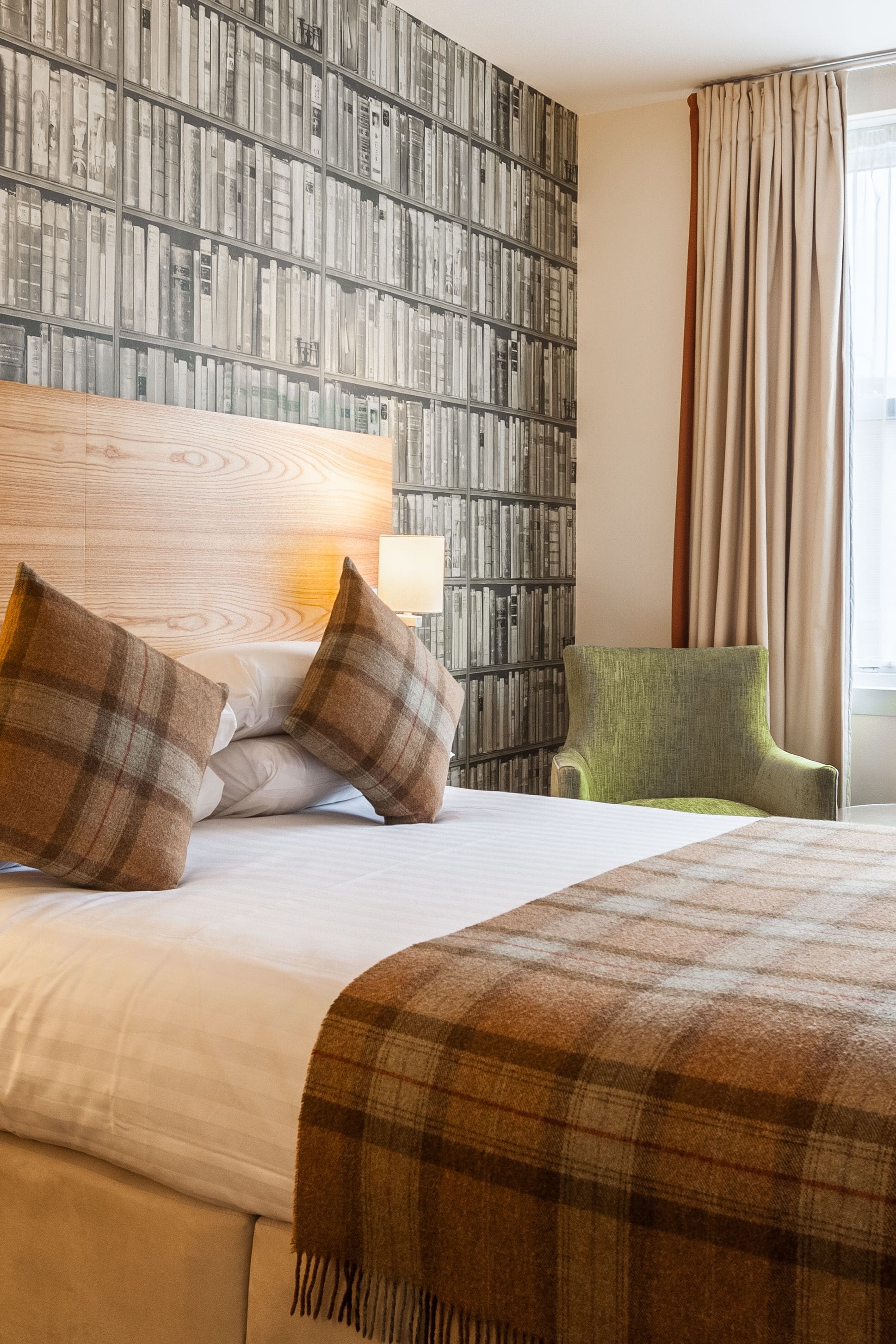 A hotel room with a large bed, a green armchair, a bookshelf, and a window with curtains.