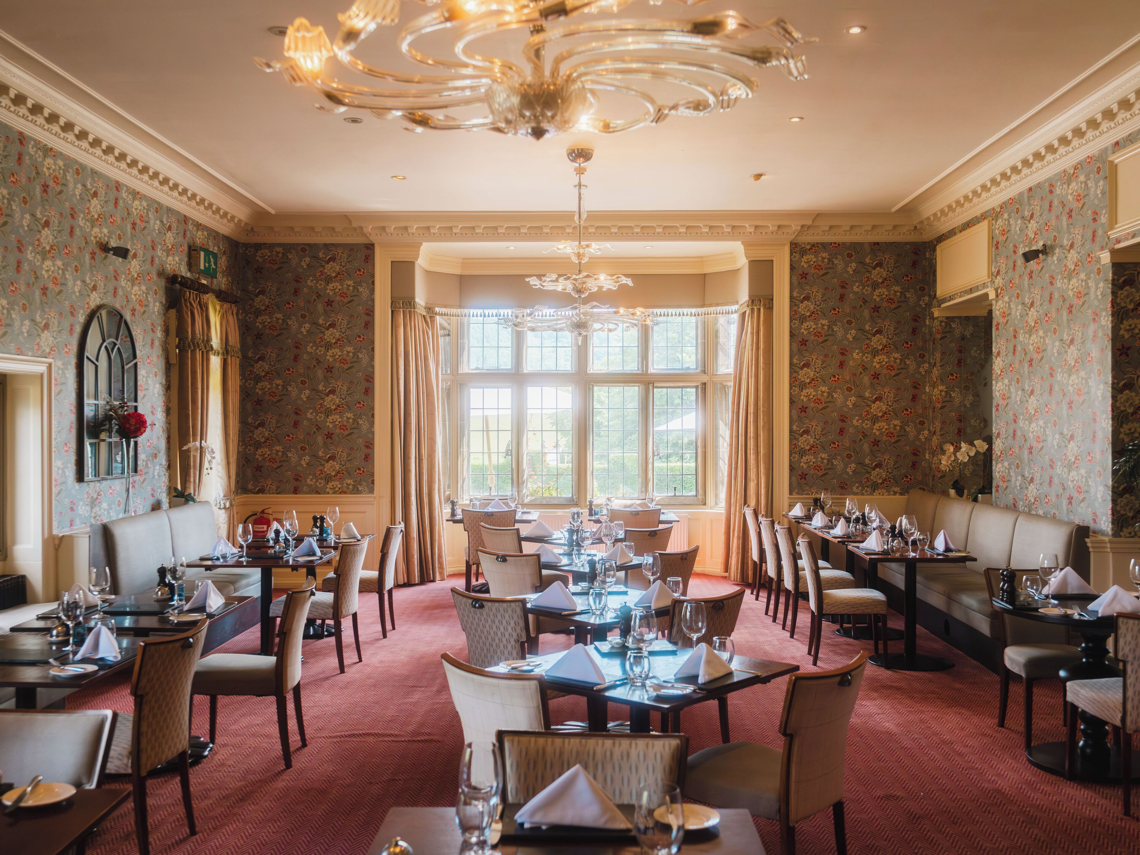 Ein elegantes Esszimmer mit mehreren Tischen und Stühlen, einer Chandelüster und floralem Wandpapier.
