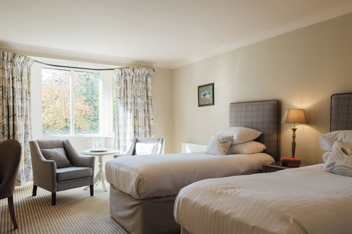 A hotel room with two beds, a chair, a small table, and a lamp.