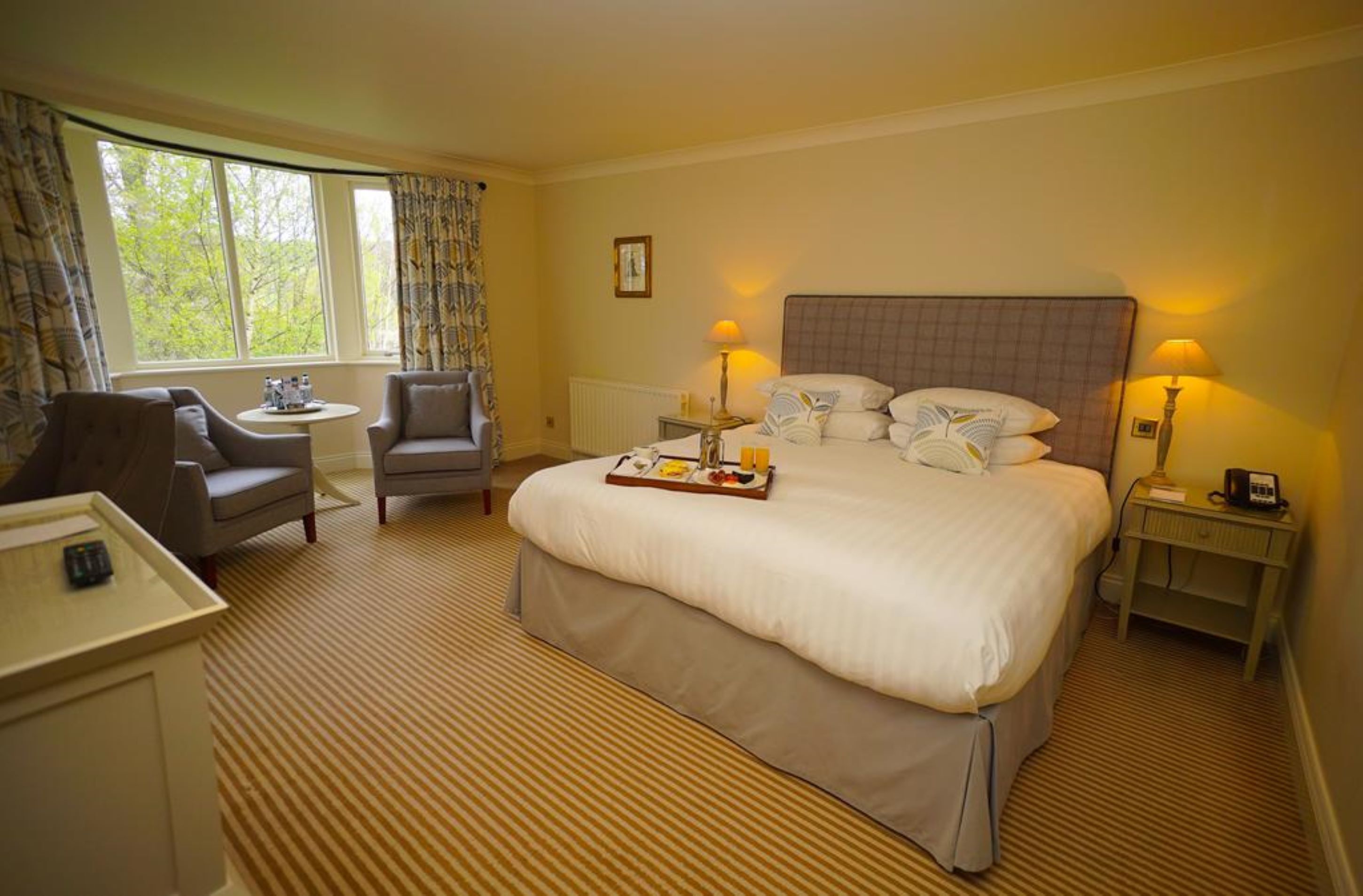 A hotel room with a large bed, two armchairs, a small table, and a bedside table with a lamp.