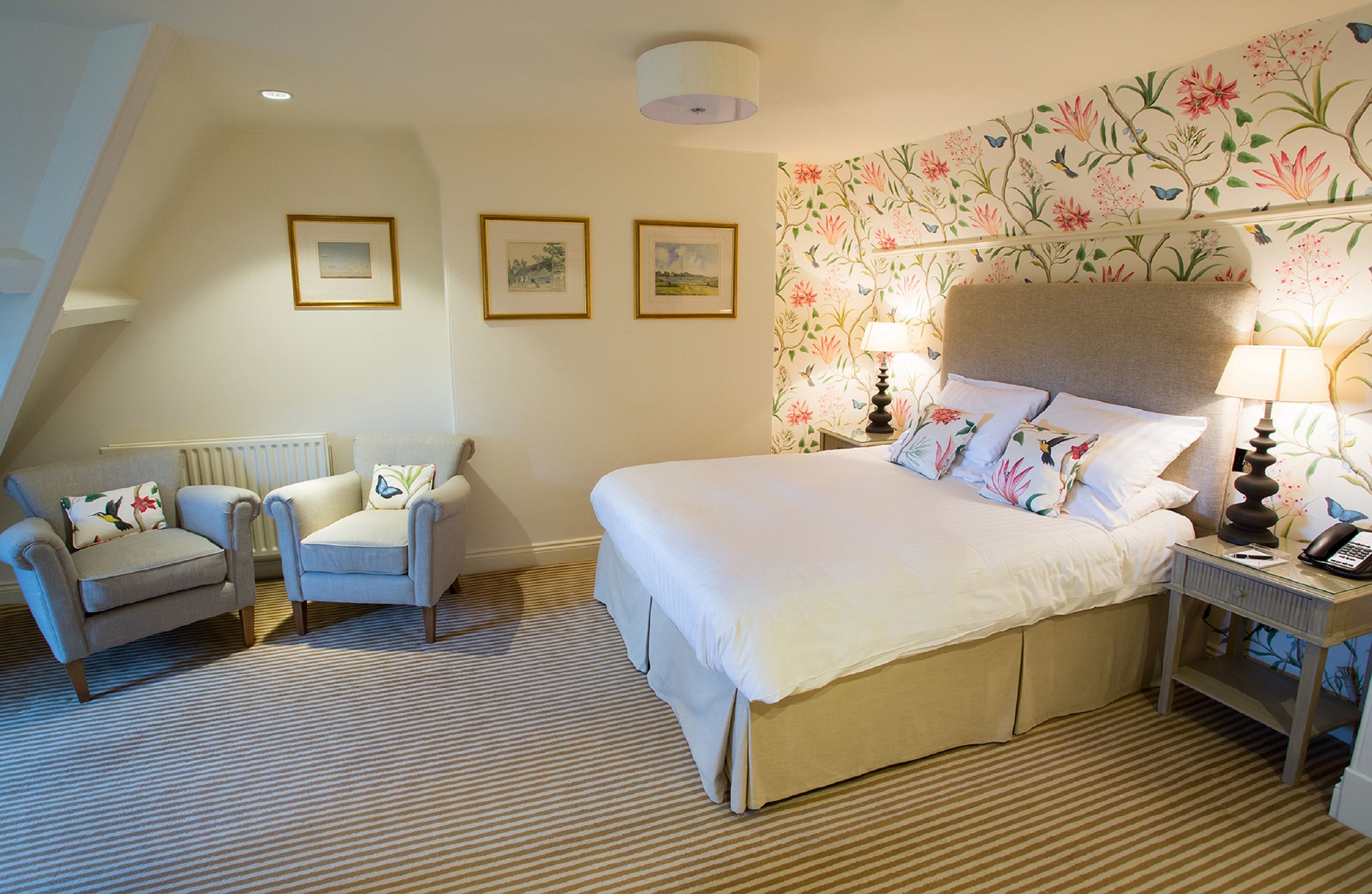 A bedroom with a large bed, two armchairs, a nightstand with a lamp, and floral wallpaper.