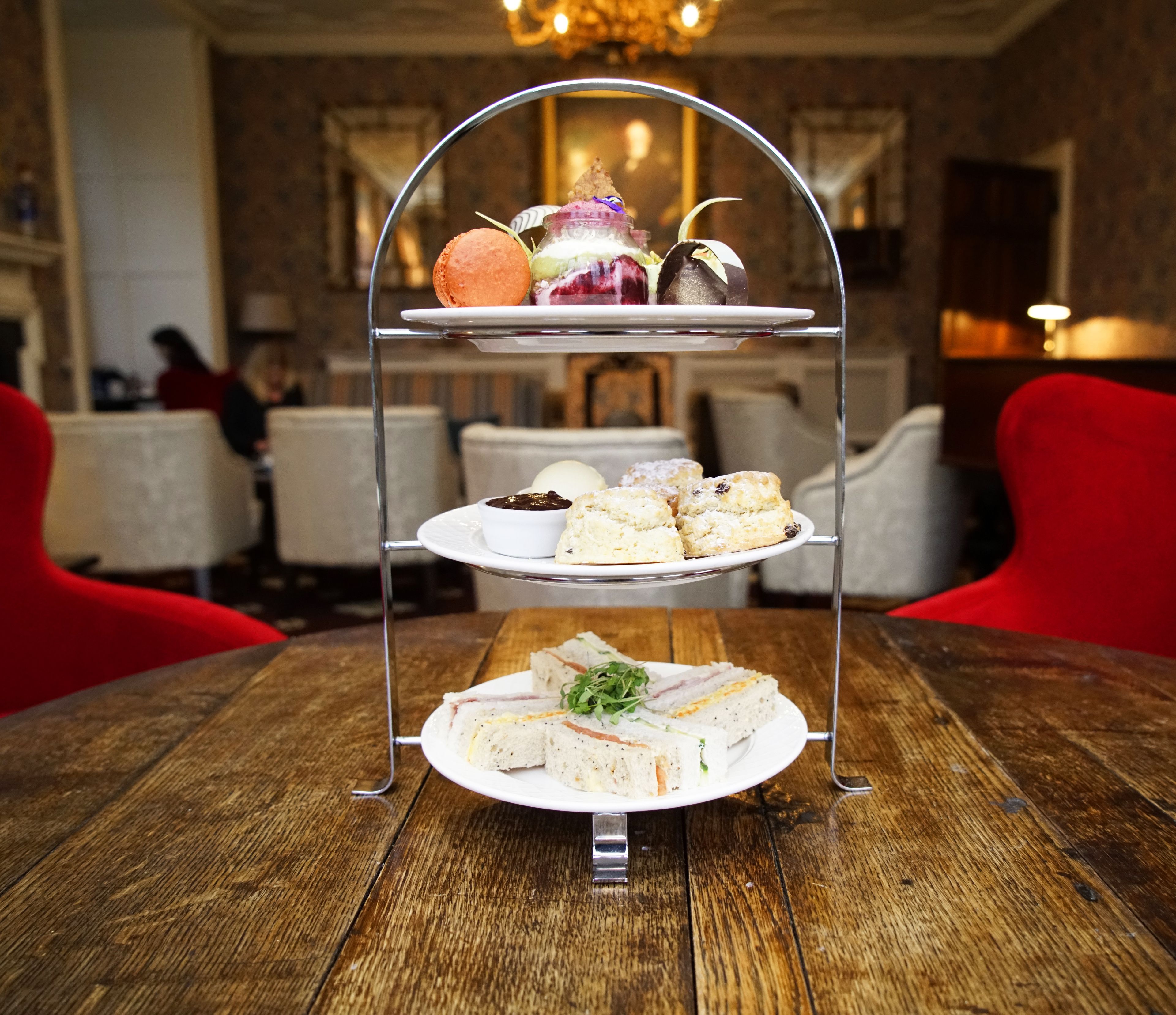 A three-tiered dessert stand with various pastries and a sandwich on a wooden table in a dining area.