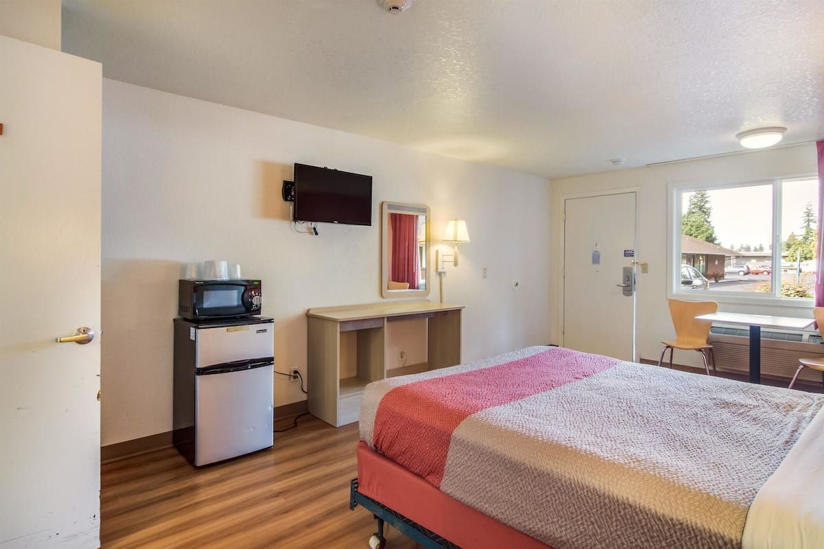 A hotel room with a bed, a desk, a microwave, a television, and a window with a view of the outside.