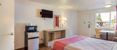 A hotel room with a bed, a desk, a microwave, a television, and a window with a view of the outside.