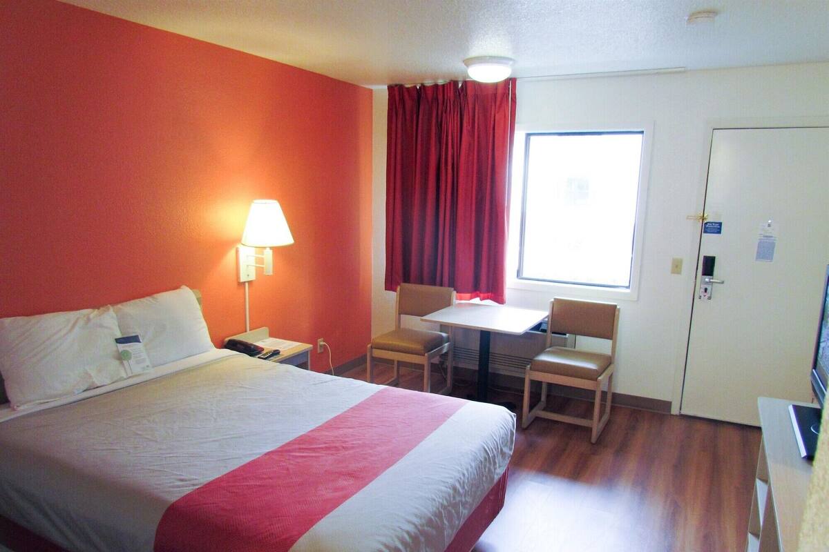 A hotel room with a bed, a desk, two chairs, a lamp, and a window with curtains.
