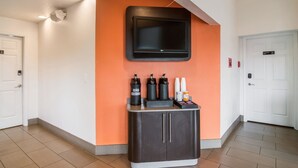 A hotel lobby with a flat-screen TV mounted on an orange wall, a minibar with beverages, and a coat rack. - Motel 6 Vancouver, WA (Vancouver)