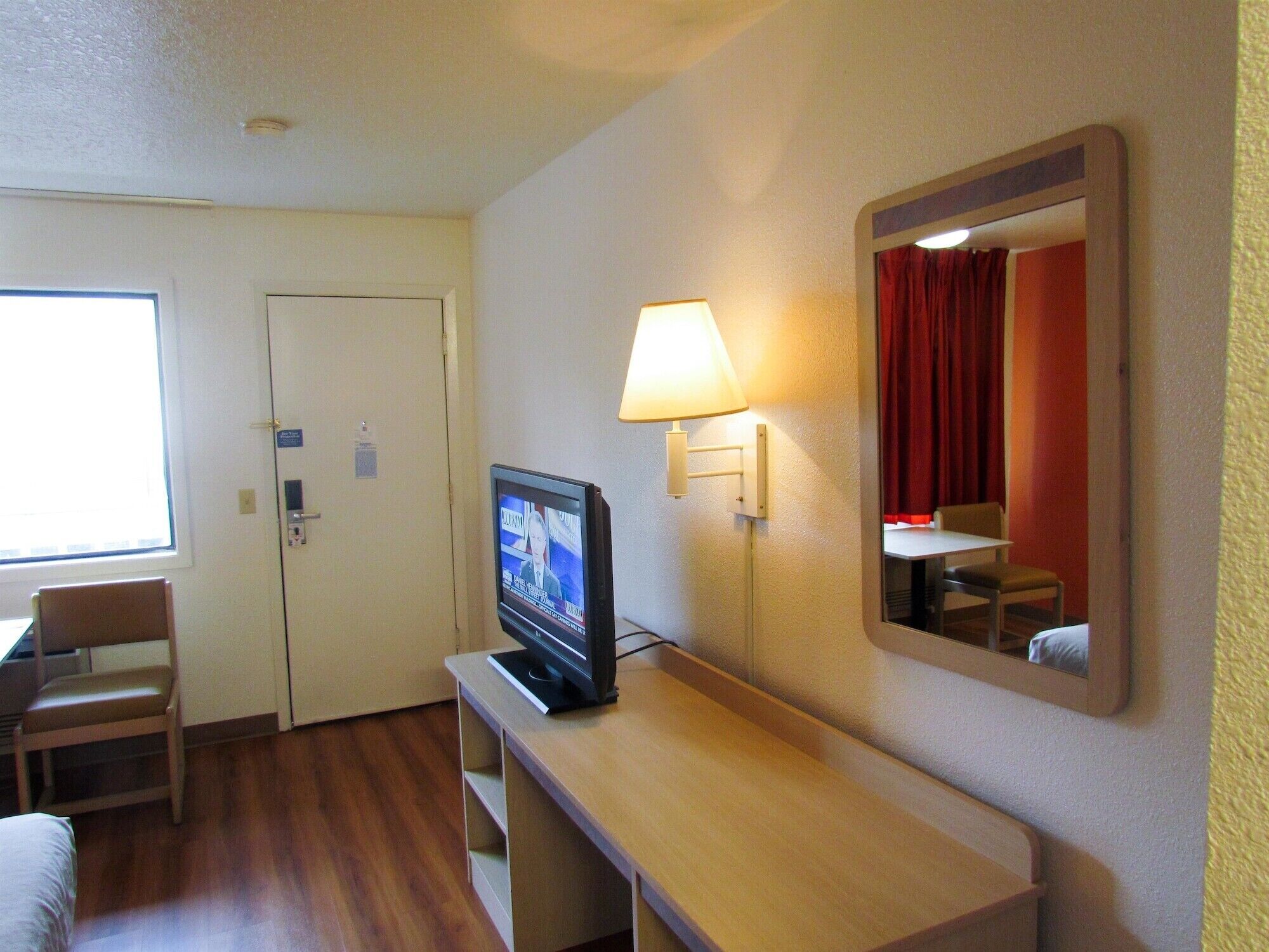 Une chambre d’hôtel avec un téléviseur posé sur un meuble en bois, un miroir au mur, une lampe, une chaise et un lit.