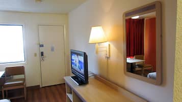 A hotel room with a television on a wooden stand, a mirror on the wall, a lamp, a chair, and a bed.