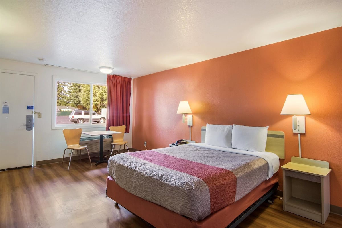 A hotel room with a bed, two bedside lamps, a desk with a chair, and a window with a view of a parking lot.
