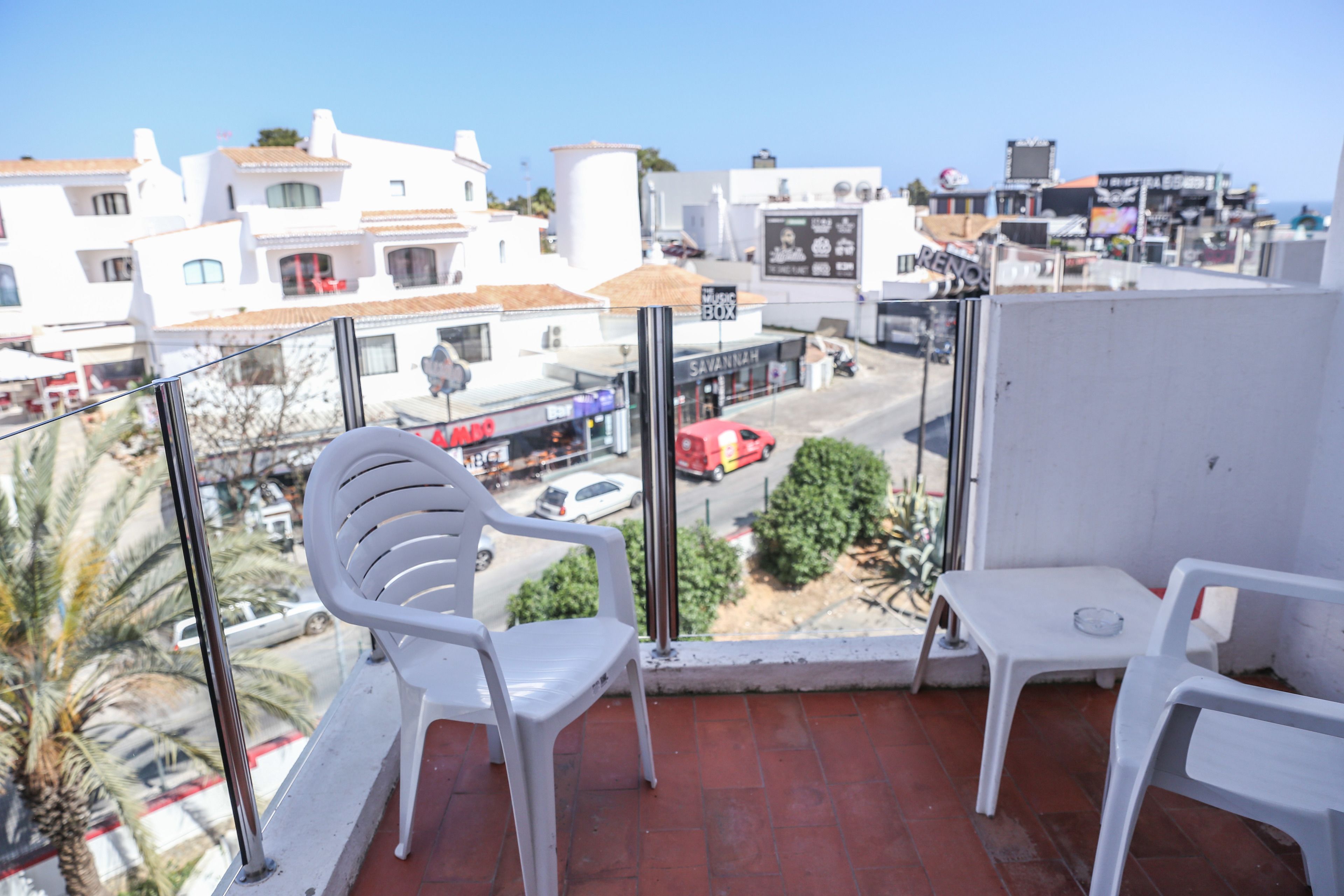 A balcony with white plastic chairs and a small table, overlooking a street with buildings and a red car.