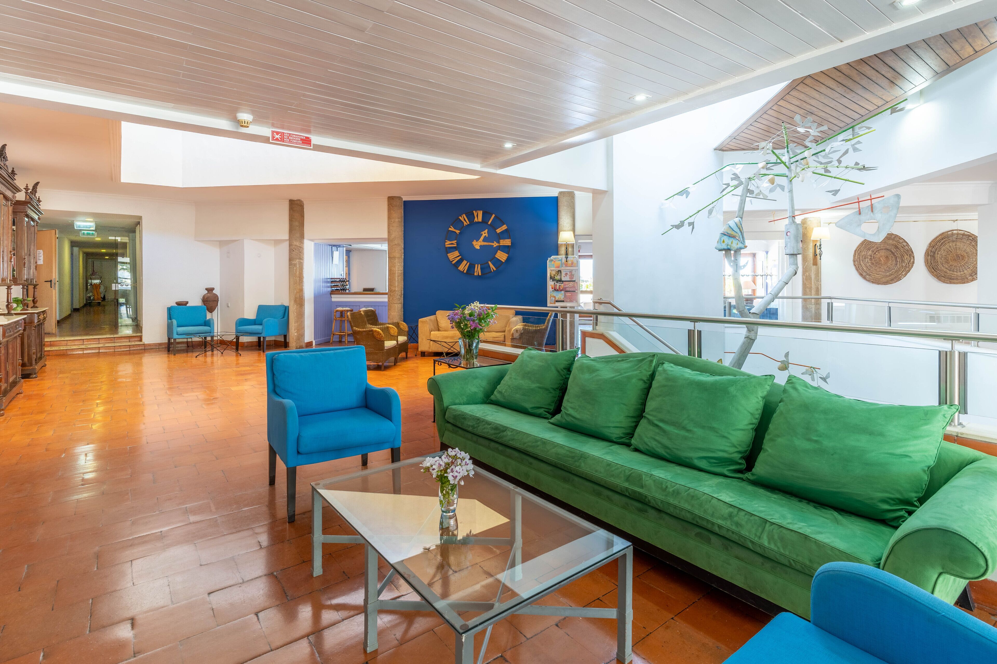 Un hall d’entrée spacieux, avec un canapé vert, des fauteuils bleus, une table basse en verre et une grande horloge murale.
