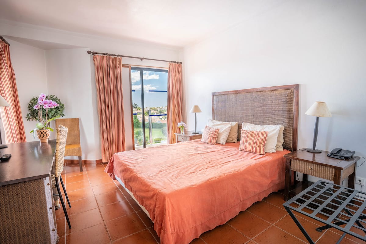 A bedroom with a large bed, a desk, a chair, a lamp, and a view of the outdoors.