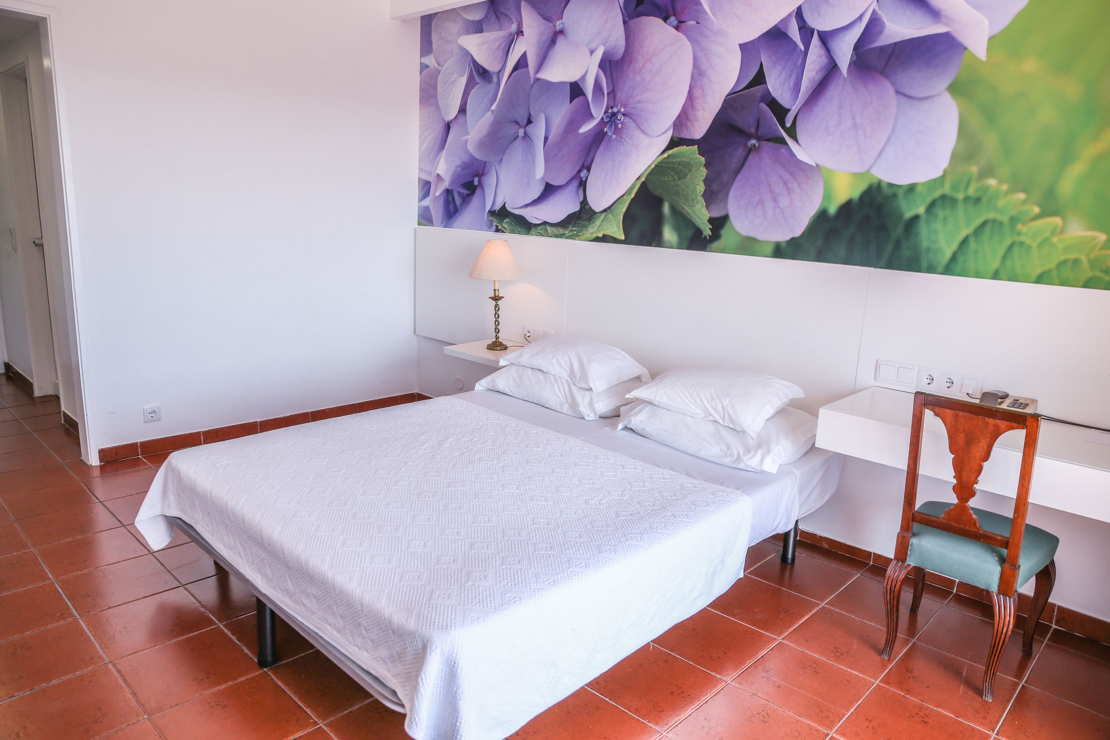 Una cama bien hecha con una colcha blanca y almohadas, una mesita de noche con una lámpara, y una silla con un cojín verde.