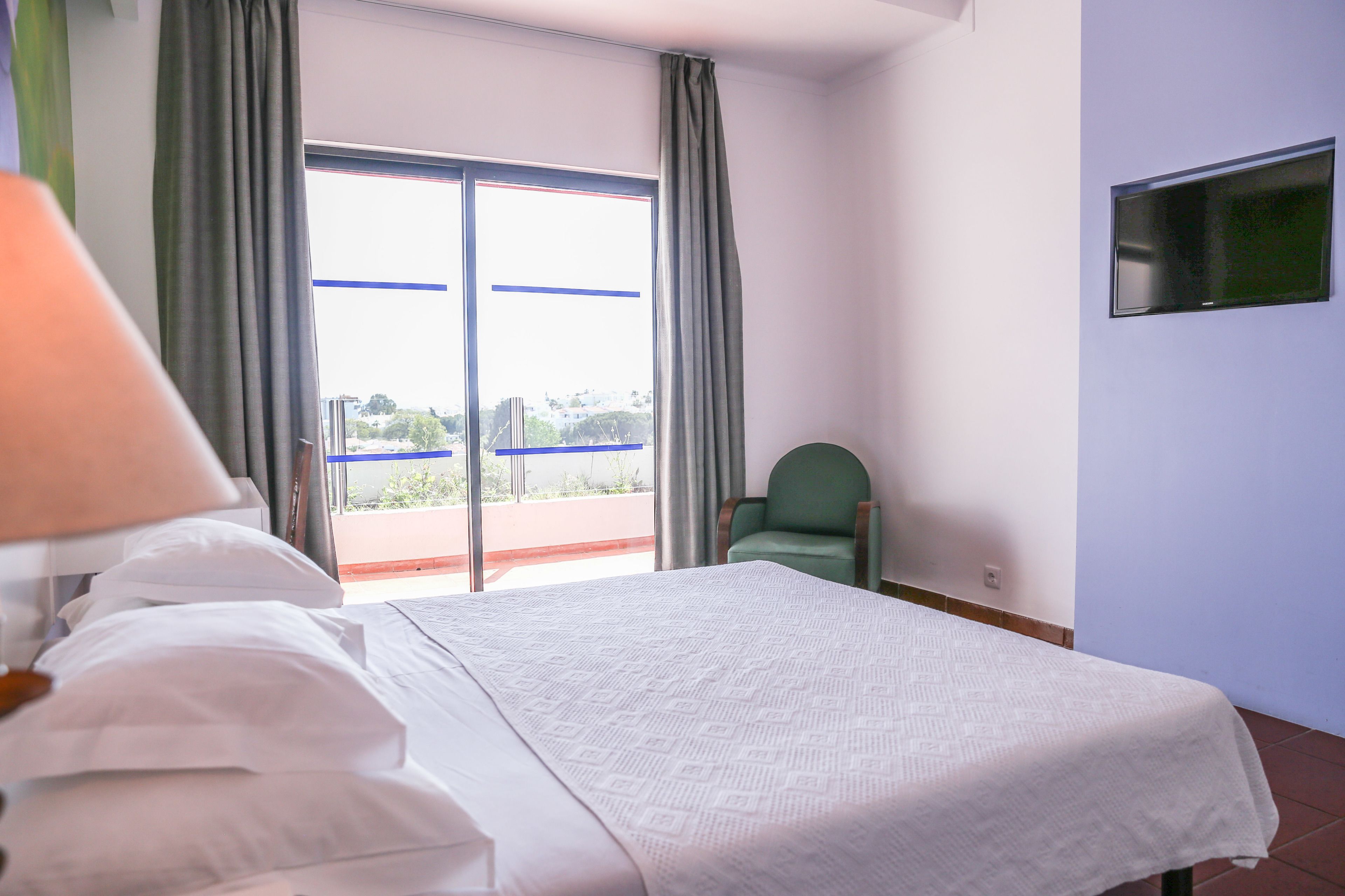 A hotel room with a bed, a chair, a TV, and a view of the outdoors.