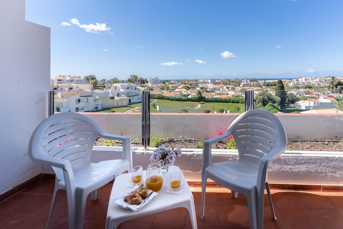 A balcony with two white plastic chairs, a small table with a plate of food, two glasses of orange juice, and a vase with flowers.