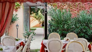 An outdoor dining area with white wicker chairs and tables, red tablecloths, and a view of a garden with plants and a pathway. - Hotel Tre Archi (Venice)