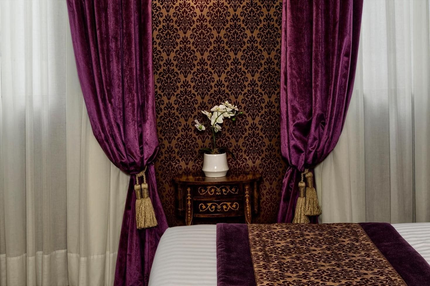 Una habitación con una cama, cortinas moradas, un papel tapiz estampado, una mesita de noche de madera y un jarrón blanco con flores.