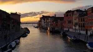 Um canal com edifícios em ambos os lados, barcos atracados na água e um pôr do sol ao fundo.