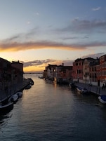 Un canale fiancheggiato da edifici, imbarcazioni ormeggiate lungo l'acqua e un tramonto sullo sfondo.