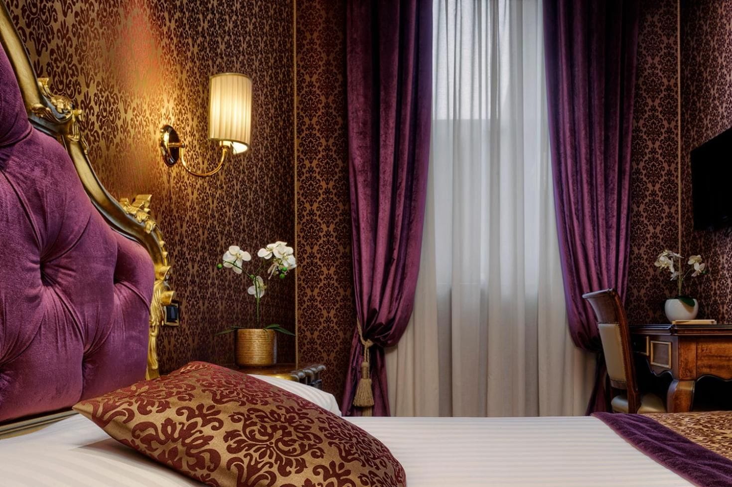Una lujosa habitación de hotel con una cama grande, un cabecero decorativo, una lámpara de pared y un ventanal con cortinas moradas.