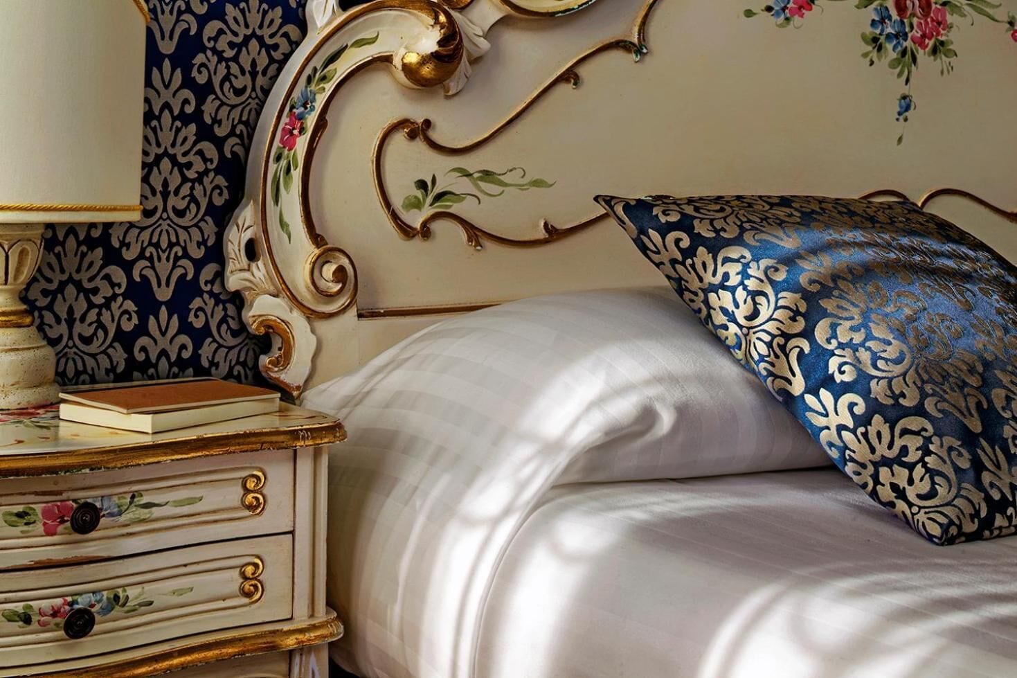 A bed with a white satin cover, a decorative headboard with floral patterns, and a nightstand with drawers and a lamp.